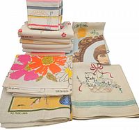 Mixed vintage linen and cotton tea towels piled showing different patterns and textures including floral and illustrated designs.