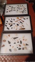 Three display cases containing over 100 prehistoric arrowheads with various shapes, colors, and sizes on white trays inside black frames.