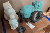 Photo showing six cat sculptures including large white cat, smaller white cat, black cat figurines, and bronze and blue-green metal cats.