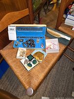 Full view of costume jewelry assortment on woven chair, showing black and white necklace, gold compact, green gem earrings and rings, silver-tone item, and other small pieces