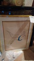 Framed art print with stylized bird in black and blue, framed in wood with glass, photographed in basement with plastic bags below.