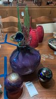 Photo showing large purple glass vase with attached green and blue glass flowers, two red ceramic vases, and two glass spheres on a wooden table