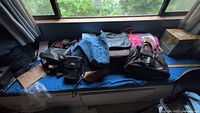 Photo of assorted handbags, totes, and denim tote lying on a bench near a window. Includes multiple black leather handbags, fabric totes in pink and purple, and a piece of denim fabric.