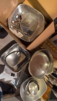 Nambe metal serving ware including various sized square and round bowls and a collection of spoons inside a cardboard box, showing overall condition and variety of pieces.