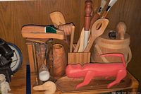 Photo showing variety of wooden utensils, red lizard sculpture, wooden whale, hourglass, carved wooden vase and part of paintbrush handles.