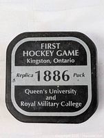 Top view of the black and white replica hockey puck with text identifying the 1886 first hockey game between Queen's University and Royal Military College in Kingston, Ontario.