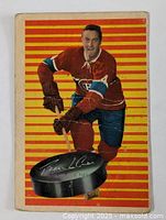 Front side of the hockey card showing Jean Beliveau in Canadiens uniform against striped orange and yellow background with printed signature on a puck illustration.