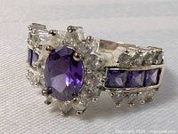Front angled view showing central oval amethyst-colored stone surrounded by clear stones and additional square amethyst stones on band.