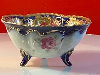 Side view of the antique footed bowl showing blue exterior, hand painted roses, and gold detailing on scalloped rim and feet.