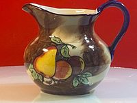 Side view of ceramic pitcher showing hand painted fruit design including a yellow pear and other fruit with green leaves on brown and cream background.