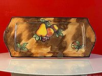 Full view of rectangular hand painted pottery tray showing mottled brown background and colorful fruit motif with handles on both ends.