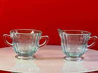 Front view of vintage blue Federal glass sugar bowl and creamer set with embossed patterns and handles, displayed against red background.