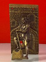 Front view of solid brass door knocker showing man figure leaning on a tool with foot on block, mounted on a rectangular backplate with arch and window textures.