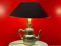 Front view of silver plated teapot base lamp with black shade against red background.