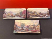 Front view of three rectangular porcelain plates featuring Thomas Kinkade landscape artistry, arranged on a dark surface against a red background.