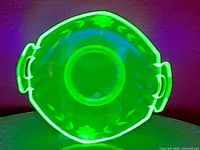 Overhead view of teal uranium glass dish glowing bright green under UV light showing octagonal shape and floral etching.