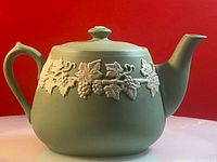 Side view of vintage green ceramic teapot showing handle, grape leaf relief band, and lid with knob.