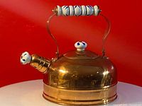Side view of vintage copper kettle showing ceramic handle and spout cap with blue floral patterns on a red background.