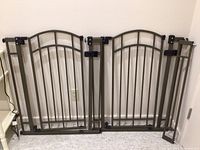 Two adjustable metal baby or pet gates with an extension piece, brown with arched tops, vertical bars, and black locking hardware, placed against a wall.