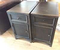 Two matching black distressed nightstands side by side showing drawer and cabinet door fronts with knobs, one drawer pull broken.