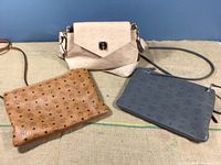Three MCM branded purses including the brown Visetos Original pouch, charcoal pouch, and Latte Beige Essential Cross Body bag arranged on a textured surface.