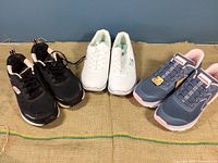 Side view of three pairs of Skechers shoes: black, white, and slate blue with pink lining on a fabric surface.