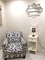 Full view including the accent chair with black and white floral damask pattern, small white side table, black lamp with wheat pattern shade, and decorative silver wall mirror
