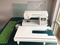 White Baby Lock Lyric sewing machine on teal surface with extension table, acrylic rulers standing behind.