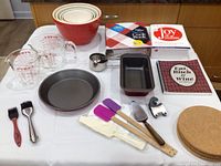 Complete set of ceramic nesting mixing bowls, glass measuring cups, baking pans, baking tools, cookbooks, and kitchen textiles on white table.