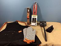 Photo showing the black apron with brown accents, striped mitt, grilling tool set box, meat claws box, grill press and stand on a beige surface against a blue background.
