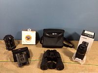 Photo showing all items arranged: Canon lens with box and white foam cradle; one pair of binoculars front and center; other binoculars beside carrying case and two black lanterns.