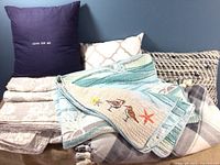 View of folded quilted bedspread, multiple pillows including a navy throw pillow and beige patterned pillows, plus plaid and sea life-themed throw blankets.