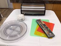 Brabantia stainless steel bread box with roll-top lid on table with lazy Susan, honey pot, and plastic cutting boards