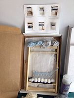Close-up of small wooden weaving loom with light blue and grey yarn woven with cardboard instruction sheet inside kit box