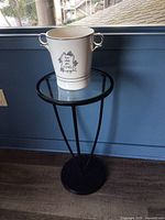 The small round table with black metal frame and glass top, shown with white ceramic ice bucket on top against a blue wall.