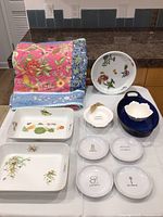 Ceramic baking dishes, punny plates, and large floral tablecloth displayed on kitchen counter.