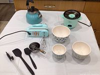 Lot of aqua kitchen appliances including KitchenAid hand mixer with beaters, kettle, and Dash cooker, plus three patterned ceramic bowls, black spatulas and metal measuring cups on white tablecloth.