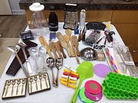 Overview of multiple kitchen utensils and gadgets including wooden spoons, graters, strainers, droppers, silicone lids, and metal straws on white tablecloth.