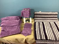 Photo showing 4 plush purple bath towels with scalloped edges stacked, 4 matching washcloths, two striped purple and gray bath mats rolled out, and a metal towel holder with two washcloths on it.
