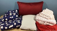 Three queen sheet sets displayed, with a red quilted throw pillow in the background. The sheets include a navy blue dog patterned flannel, a beige gingham cotton set, and a cream with red geometric patterned set.