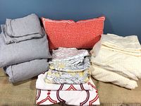 Two sets of queen sheets and pillowcases folded and arranged on fabric surface including yellow cotton damask sheets, grey UGG flannel sheets and various folded sheets with floral and geometric prints.