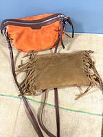 Orange suede Rag & Bone purse with brown leather strap, and brown suede UGG purse with fringe detail and long strap, both laid on a surface.