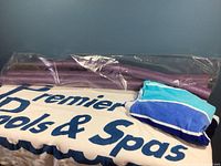 Three new purple pool noodles wrapped in plastic and three swim towels of varying blue shades placed on a towel with 'Premier Pools & Spas' text.