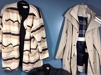 Three jackets hung on a blue wall: a cream and brown striped plush jacket, a tan raincoat with plaid inner lining, and a black quilted nylon jacket.