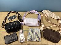 Three small leather purses in black, pale lilac, and brown colors with multiple fabric straps and a pom pom accessory on brown purse. Additional black pouch and packaged straps also visible.