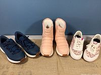 Three pairs of Kizik shoes shown side by side: navy athletic shoes, pink boots, and white floral sneakers.