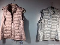 Two sleeveless quilted vests hanging on wall - pale grey with lavender tint and light blue, both size Large by 32 Heat