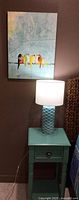 Full view of the lamp on a turquoise side table with the bird painting hanging on the wall above it.