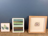 Set of three wall art pieces arranged side by side against blue wall on burlap surface
