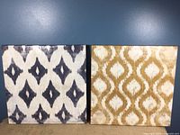 Two square 24x24 inch wall artworks with patterned designs, one gray/white diamond motif and one gold/white ornamental motif.